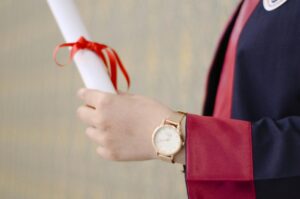 Close-up of a graduate holding a rolled diploma tied with a red ribbon, wearing a formal graduation gown.