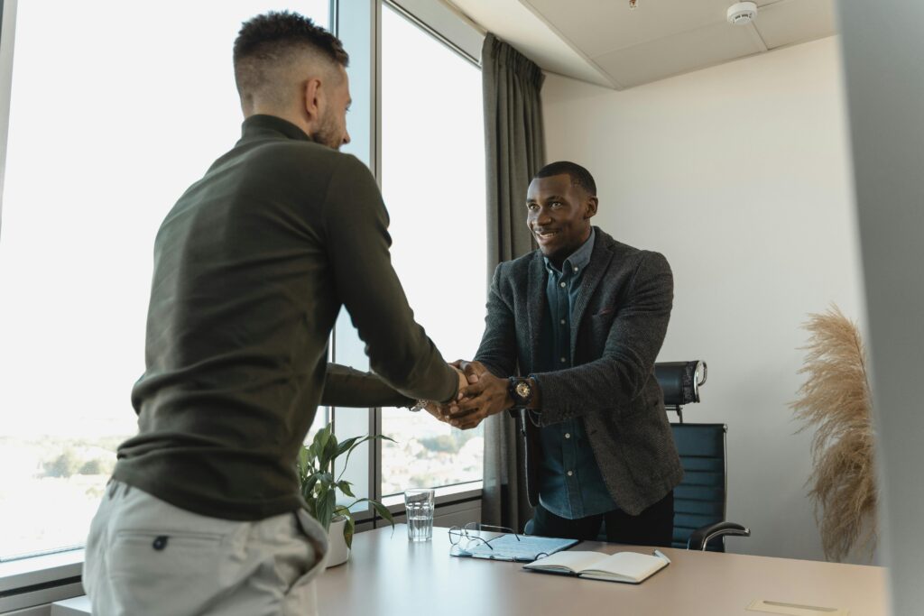 Two businessmen shaking hands across a desk in a modern office, symbolizing partnership and professional agreement.